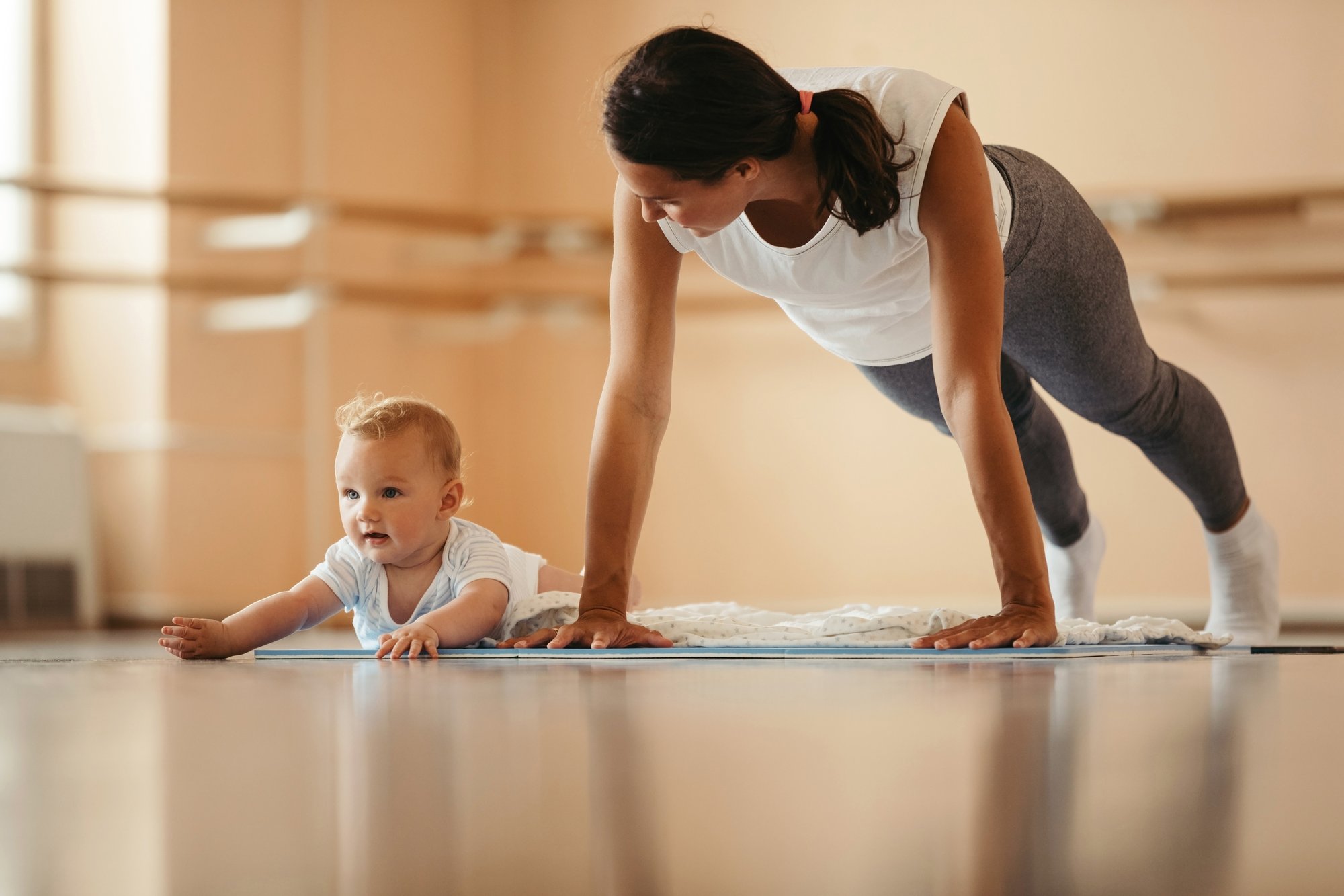 Mutter in Plank-Position auf der Yogamatte, ihr Baby krabbelt neben ihr